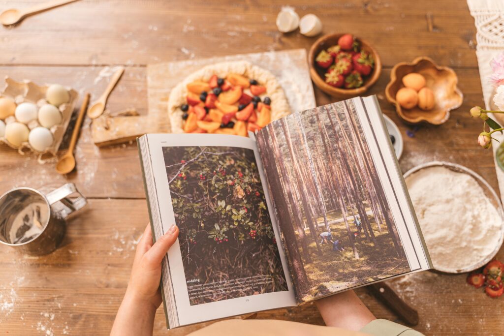 Top view of a person holding a book surrounded by baking ingredients and fresh fruits on a wooden table.